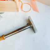 A metal and wood hammer with a rectangular head rests on a light surface near a SheerID Wood Box Brand box, an orange cord, and a piece of orange material.