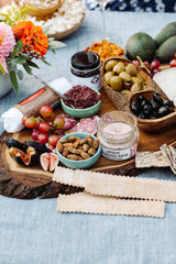 A wooden platter holds green grapes, figs, crackers, almond pâté, nuts, salami, jam, cheese, and Sun Dried Balsamic Black Olives on a blue tablecloth with flowers nearby.