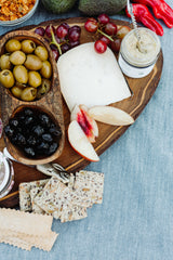 A wooden platter with Sun Dried Balsamic Black Olives, green olives, cheese, red grapes, apple slices, seed crackers, a jar of spread and jam, set on a blue cloth.