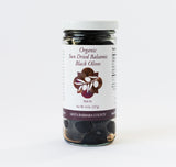 Sun Dried Balsamic Black Olives in an 8 oz glass jar, organic and sun dried in balsamic vinegar. Jar features a white label with brown and purple text plus an olive branch illustration.