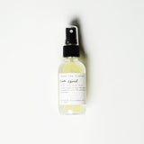 A clear spray bottle labeled Sun Spirit Cooling Aloe Mist with a black nozzle contains yellow liquid. The label notes it’s a gentle aloe and essential oils mist for post-sun relief, handcrafted in California. The bottle is on a white background.