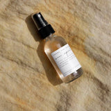 The Sun Spirit Cooling Aloe Mist, in a small clear spray bottle with a black nozzle and white label, sits on sunlit beige fabric, its shadows and light forming soft patterns across the surface.