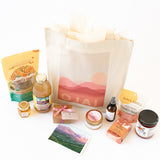 The Ojai Gift Tote includes a canvas bag with a pink sunset and Ojai text, surrounded by local treats—snacks, small honey jar, juice, candle, skincare items, a pink-wrapped box, and a mountain scene postcard.