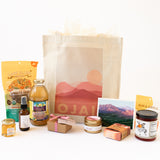 The Ojai Gift Tote features a canvas bag with a pink mountain “OJAI” design, plus gourmet snacks, juice, soap bars, honey jar, local treats, spray bottle, and a scenic postcard—offering a warm Ojai welcome.