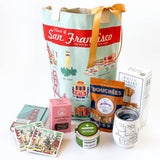 The San Francisco Gift is a city-themed gift bag featuring local snacks, cookies, coasters, a chocolate box, canister, and rustic bakery box, all neatly arranged and finished with a gold ribbon tied to the handle.