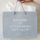 A hand holds the Welcome to Santa Barbara Grey Gift Tote, featuring "Welcome to Santa Barbara 34.4° N / 119.7° W" in white letters. White tissue paper peeks out against a textured, off-white wall. Eco-friendly design.