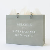 The Welcome to Santa Barbara Grey Gift Tote is an eco-friendly bag made from recycled paper, featuring gray rope handles, white tissue paper, and "WELCOME TO SANTA BARBARA 34.4° N / 119.7° W" in white capital letters.