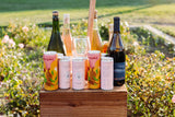 A wooden crate outdoors holds colorful cans of Ysidro Grapefruit Sake Spritz, wine bottles, and glasses, surrounded by greenery and flowers in soft sunlight.