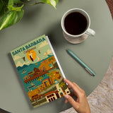 Hand holding a Santa Barbara Scene Journal on a table with coffee, pen, plant, and 60 lined pages inside.