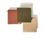 Three closed Square Linen Blank Notebooks in brown, green, and beige, each with a ribbon bookmark and fabric cover.
