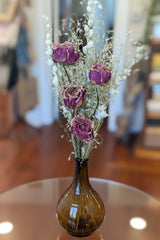 The California Dried Floral Arrangement with purple roses and white foliage sits on a reflective table in a unique vase.