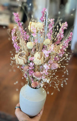 A hand holds a California Dried Floral Arrangement of pink and beige flowers against a blurred background.