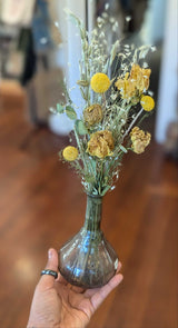 A hand holds the California Dried Floral Arrangement indoors on a wooden floor.