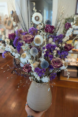 A hand holds the California Dried Floral Arrangement with dried flowers in purple, white, and beige tones.