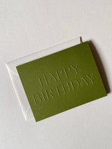 Olive green Happy Birthday Note Card, embossed with HAPPY BIRTHDAY in capital letters, resting on a matching envelope.