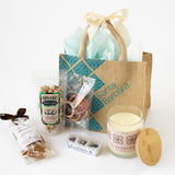 The American Riviera Tote gift set features a jute bag with cream bow, wooden-lid candle, organic pistachios, dried orange slices, assorted caramels, and chocolates—local treats inspired by Santa Barbara charm.