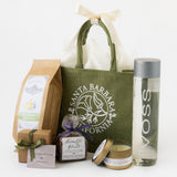 The An Elegant Escape Santa Barbara Gift Tote is surrounded by a VOSS water bottle, travel candle, small soap bar, fine chocolate truffles, a gift box with bow, lavender sachet, and sea salt olive oil crackers.