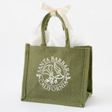 The An Elegant Escape Santa Barbara Gift Tote is a green jute bag featuring "Santa Barbara California" and a white bird design. It includes white-ribbon-tied handles and tissue paper—ideal for gifting with travel candles or fine chocolate truffles.