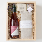 An open wood gift box with a bottle of Morét-Brealynn rosé, a Dead Sea Minerals soap bar, a Riviera Gardens bath bomb, and a linen lavender eye pillow, arranged on cream crinkle paper with dried lavender sprigs.