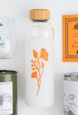 The California Poppy Water Bottle features clear glass, a bamboo lid, and a white sleeve with an orange poppy design. It’s pictured with a candle, glass, book, and tumbler.