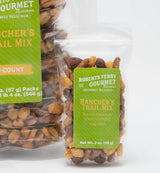 A clear plastic bag labeled California Rancher's Trail Mix contains gourmet nuts, corn nuts, and snacks. The Roberts Ferry Gourmet label and Net Wt. 2 oz. (59 g) are visible, with a larger pack partially visible in the background.