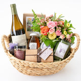 The Calle Elegante Gift Basket (Seasonal) features a wine bottle, floral bath tea, honey jar, candles, chocolate truffles, gift boxes, and a colorful flower bouquet—beautifully arranged for Santa Barbara gifts.