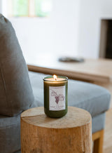A lit candle in a green glass jar sits on a wooden stump by a gray sofa in a modern living room. The Calle Elegante Gift Basket (Seasonal) makes the perfect Santa Barbara gift—pair it with chocolate truffles for extra thoughtfulness.