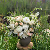 The Grande, a neutral ceramic pot, features a lush mix of sustainably grown white and pale peach flowers with greenery, displayed outdoors among tall grass and soft-focus background greens.
