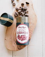 A cylindrical container labeled Cheerful Wishes Holiday Gift Box sits open on a wooden board, with chocolate-covered cherries scattered around it and the lid set aside.