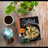 The Crafting a Meal Cookbook rests on a rustic wooden table next to coffee, a sugar bowl with a wooden spoon, and a potted plant—ideal for discovering Mediterranean-inspired Santa Barbara recipes.