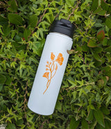 The Custom Logo Sequoia Stainless Tumbler, featuring an orange floral design on white stainless steel, rests among green leafy plants and vines.