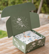 The "Customize Your Own Mailer Gift Box," featuring a green box with white floral designs and a builder-themed inner box adorned with colorful botanicals, is displayed outdoors on a wooden surface.