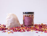 A reusable glass jar labeled “Floral Bath Tea” sits by a drawstring sachet and fragrant dried organic florals, all arranged on a white surface with a plain background, inviting you to unwind with Floral Bath Tea.