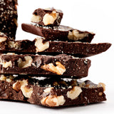 A close-up of stacked pieces of Ginger Walnut Dark Chocolate Bark, filled with large walnut chunks, set on a wooden table against a white background.