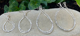 Four pairs of Gold Pear Shaped Hammered Hoops from Ocean Jewel displayed on stone with green leaves in the background.