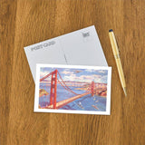 A Golden Gate Bridge Postcard, gold pen, and blank postcard arranged on a wooden surface.