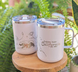 Two white insulated camp mugs with clear lids sit on a wooden stand. One showcases a bear and flowers; the other is the Golden State California Insulated Camp Mug reading “Golden State California est. 1850.” Lush ferns blur behind them.