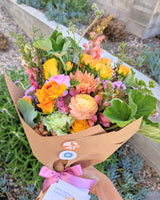 A hand-tied bouquet of fresh flowers with a mix of orange, yellow, pink, and green colors, elegantly wrapped in brown paper with a pink ribbon.
