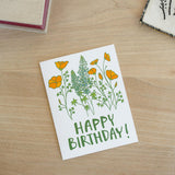 The Happy Birthday California Poppy Note Card rests on a wooden surface, displaying orange and green flowers with "HAPPY BIRTHDAY!" in green. Books and fabric peek from the corners.