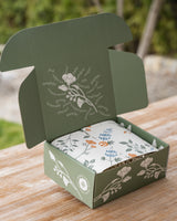 The Happy SB Treats Gift Box—a green cardboard box with floral designs and white text inside the lid—holds Santa Barbara snacks like organic popcorn and handcrafted chocolate truffles, displayed on a wooden outdoor surface.