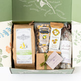 The Happy SB Treats Gift Box includes Santa Barbara snacks such as sea salt & olive oil popcorn, organic pistachios, a macadamia nut bar, sea salt caramels, and a small brown gift box tied with a green ribbon.