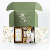 The Happy SB Treats Gift Box features a green floral box with an open lid, showcasing coffee, nuts, organic popcorn, a snack bar, and a small wrapped gift—all attractively arranged inside.