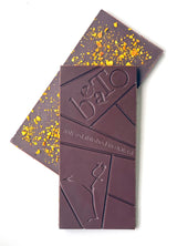 Two Happy Valley Chocolate Bars with Orange Oil & Ojai Pixie Dust are stacked on a white background: the top bar is engraved with "beTa anti established in ojai, ca" and a stylized figure; the bottom bar is topped with golden crumbs.