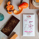 A Happy Valley Chocolate Bar with Orange Oil & Ojai Pixie Dust is artfully arranged with Ojai Pixie mandarin slices, candied orange peels, a turquoise stone, and a chocolate bar on a light surface, viewed from above.