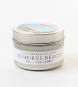 A 3oz silver tin labeled "Hendry's Beach Travel Candle" from Santa Barbara Company, with an abstract beach and ocean illustration on the lid.