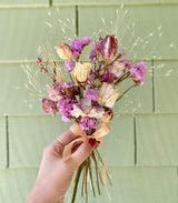 A hand holds the Garden Dried Floral Bouquet, tied with a peach ribbon, in front of a green shiplap wall.