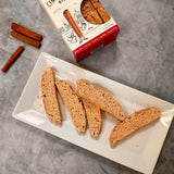 Four biscotti on a plate beside a Cinnamon Toast Biscotti box and two cinnamon sticks on a gray surface.