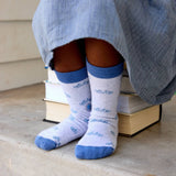 A child in a blue dress sits on books, wearing Kids Socks Gray Bicycles—organic socks with blue trim.