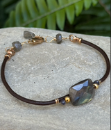 The Labradorite Reflection Bracelet features a large iridescent stone, gold accents, and gray beads on a stone surface.