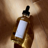 A glass dropper bottle of Lavender Body & Bath Oil rests on wood, casting a shadow. The clear oil gleams in the light, showcasing its minimalist white front label.